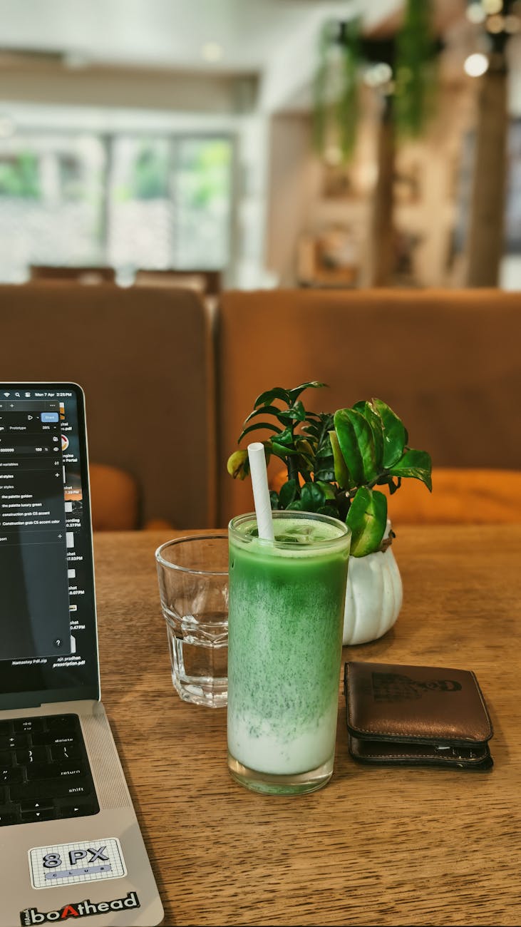 A laptop, matcha latte, and a small plant on a wood cafe table in warm window light.
