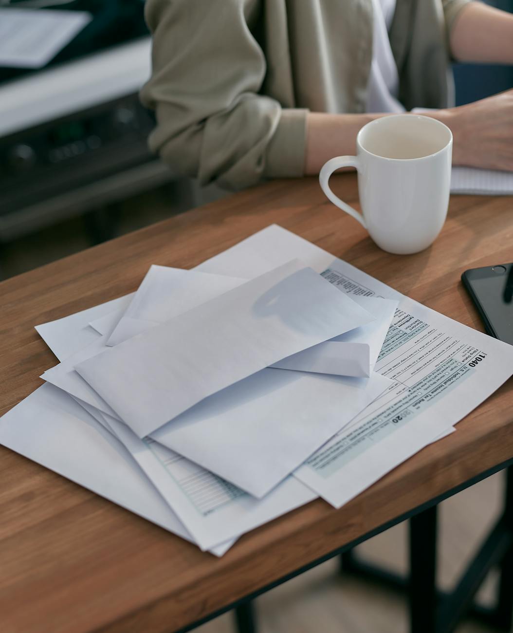 A stack of opened envelopes and a 1040 tax form spread across a kitchen table beside a coffee mug.