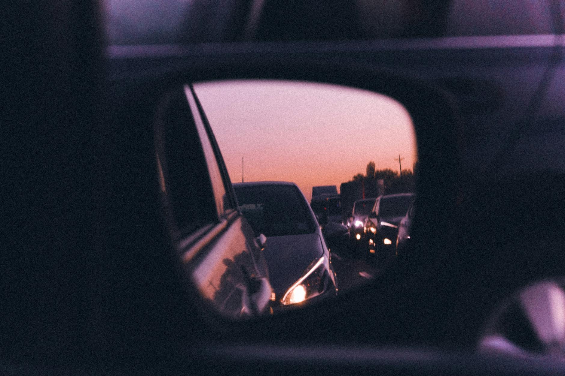 View through the side mirror of a car stuck in highway traffic at sunset, brake lights ahead.