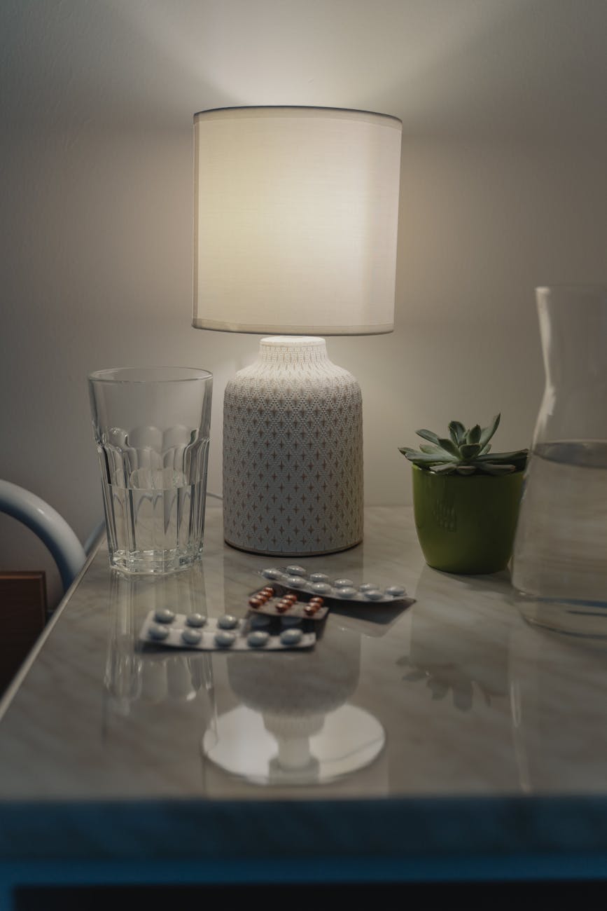 A bedside table at night with a lamp, a glass of water, and a blister pack of pills laid out.