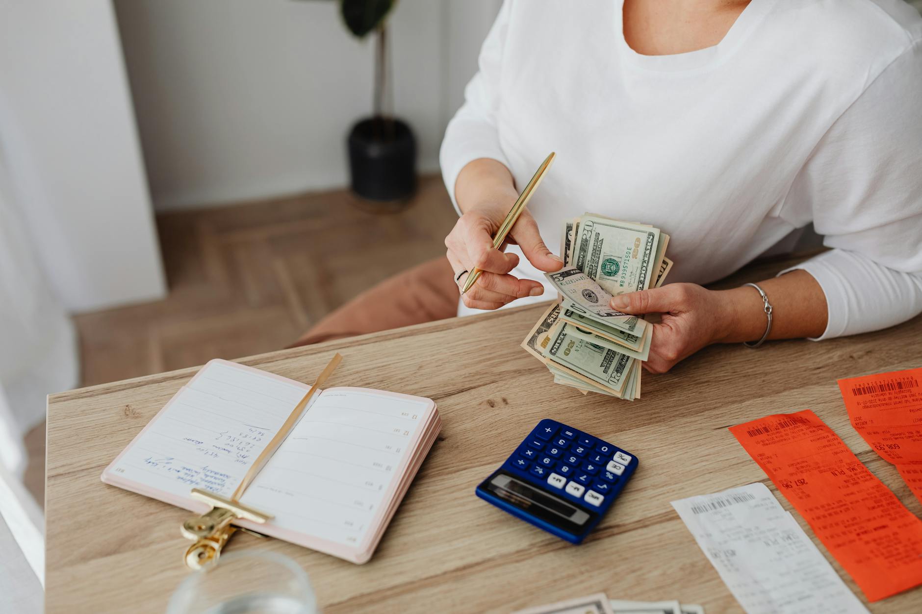 A wooden table with a notebook, calculator, receipts, and cash being counted by hand.